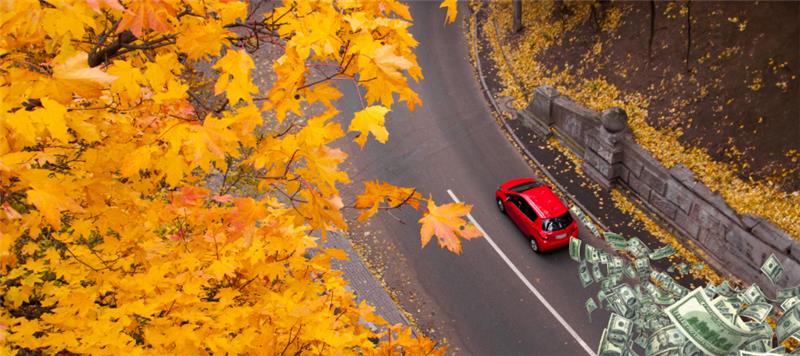 a red car drives on a fall road while money flitters in the breeze