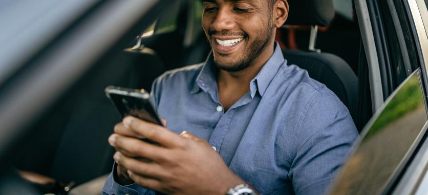A smiling man is looking at his phone in the driver's seat of a car.