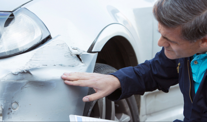 A man with a clipboard squatting to inspect a dent on the front of a car