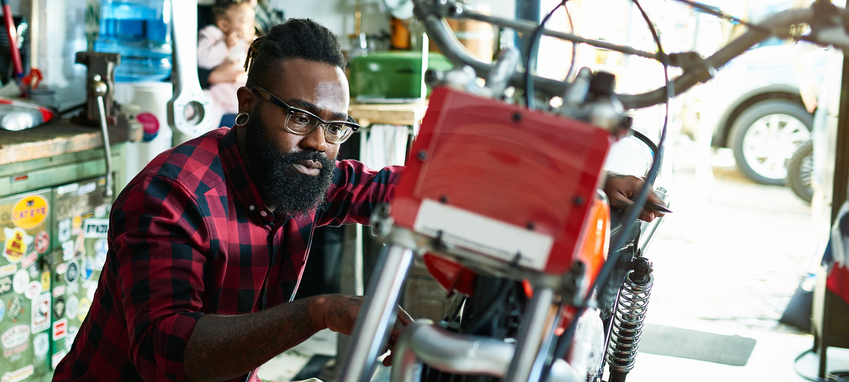 Bearded man fixing motorcycle in motorcycle workshop, woman with baby girl in background.
