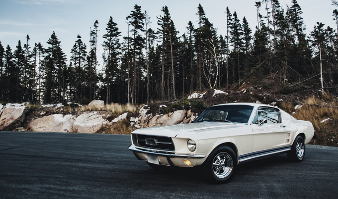 Classic car (Mustang) parked on a road in a forest/mountain landscape.