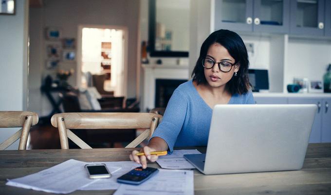 Woman with glasses on laptop entering something into a calculator.