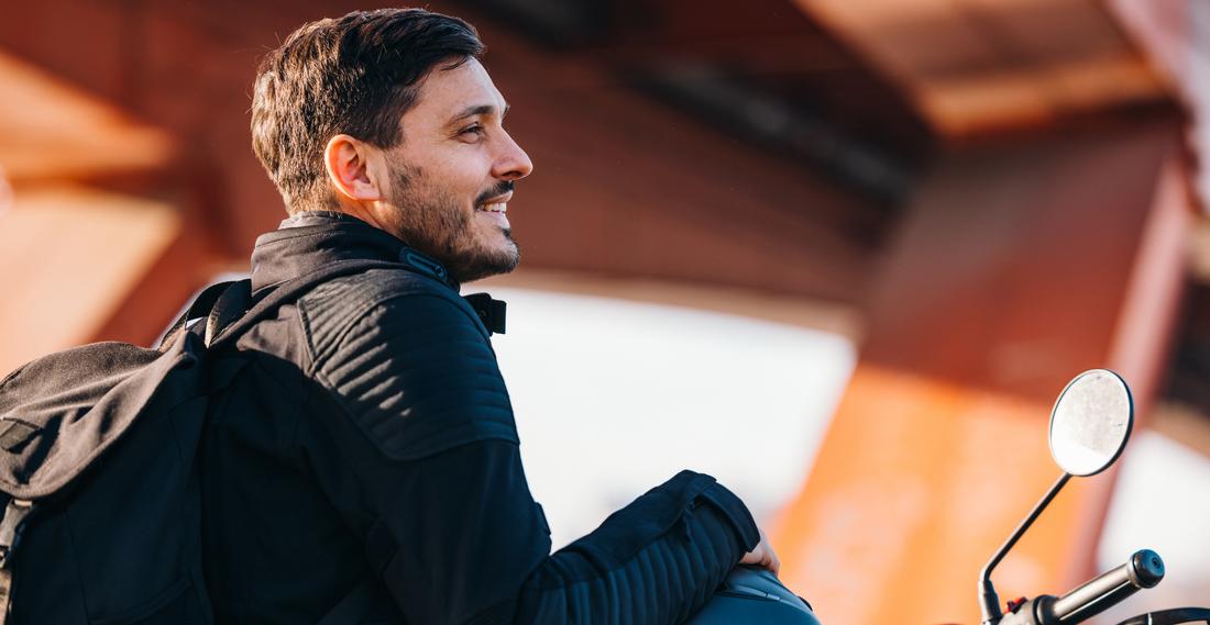 A side view of a smiling man in a black jacket and black backpack sitting on a motorcycle, holding a black helmet on his lap, with an orange steel bridge in the background.