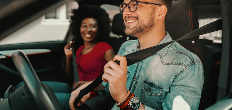 A man and a woman are smiling in a car putting on heir seatbelts.
