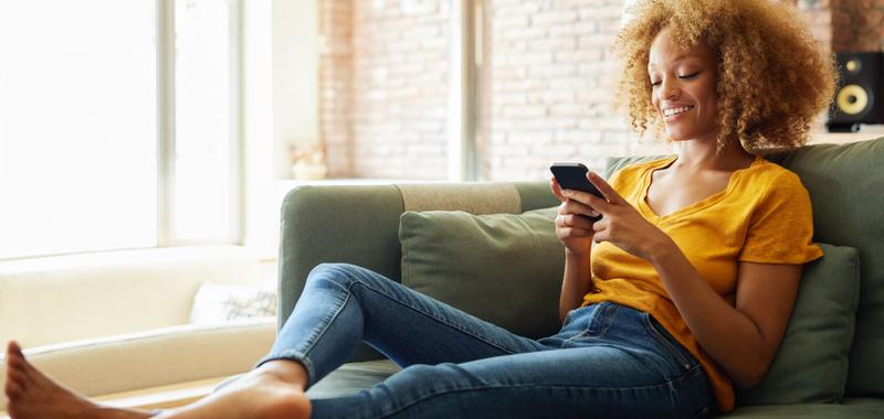 woman sitting on couch barefoot smiling while looking at her phone 