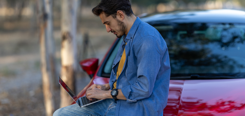 A man uses a laptop while sitting on a car hood