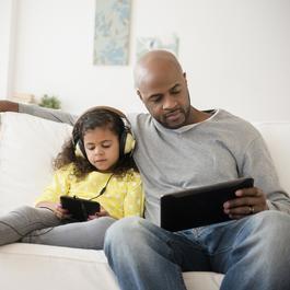 Dad and daughter sitting together on the couch, playing on electronic devices.