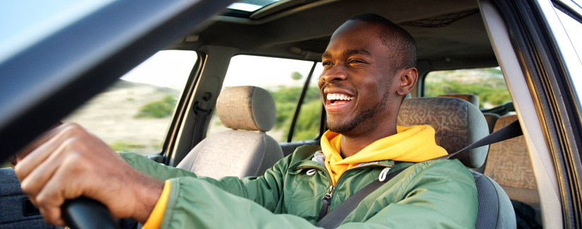 A smiling young man is sitting in the driver's seat of a car with his hands on the steering wheel.