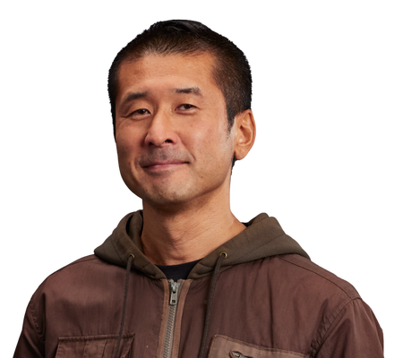 photo of an ethnic young male smiling wearing a brown sweater and gray pants with hands in pocket