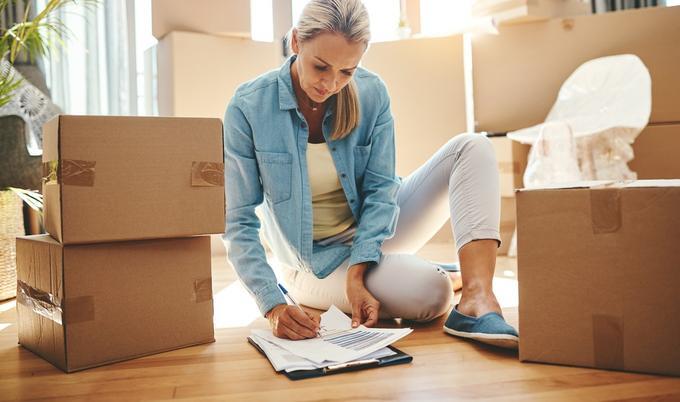 Woman on floor writing on papers on a clipboard with cardboard moving boxes all around her.