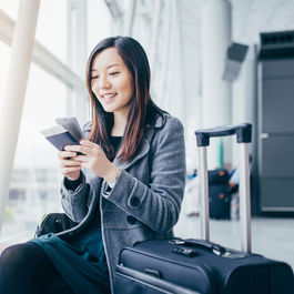 Young woman sitting at airport with her passport and phone in her hand, next to her luggage and a camera on the seat next to her.
