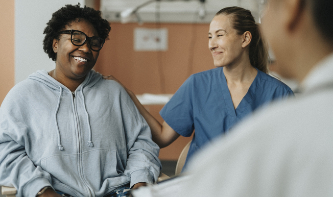 A smiling woman in a hoodie is in a hospital room with a smiling nurse who has her hand on her shoulder.