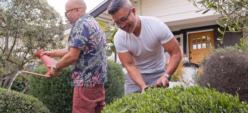Two men gardening in front of a house. The man in the foreground is smiling and trimming a bush. The man in the background is watering a bush with a red watering can.