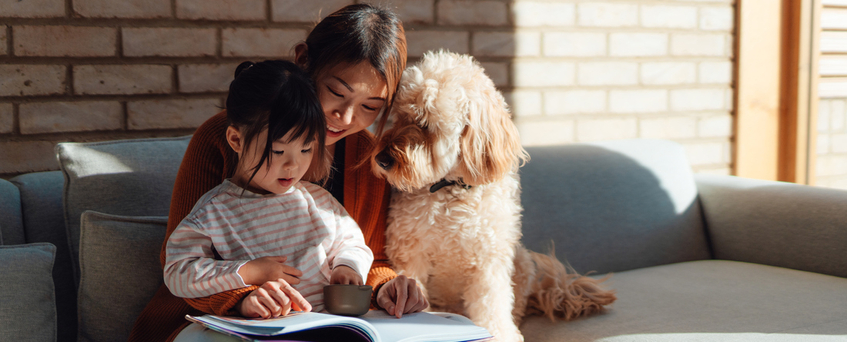 Toddler daughter sitting on her mother's lap, reading a book, enjoying story time, relaxing on the sofa at home with their dog.