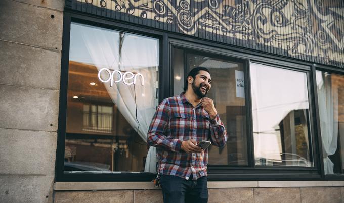 A smiling man standing in front of a restaurant while holding his smartphone