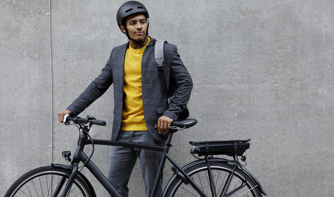 A young man in a helmet is standing behind a black eBike with a concrete wall in the background.