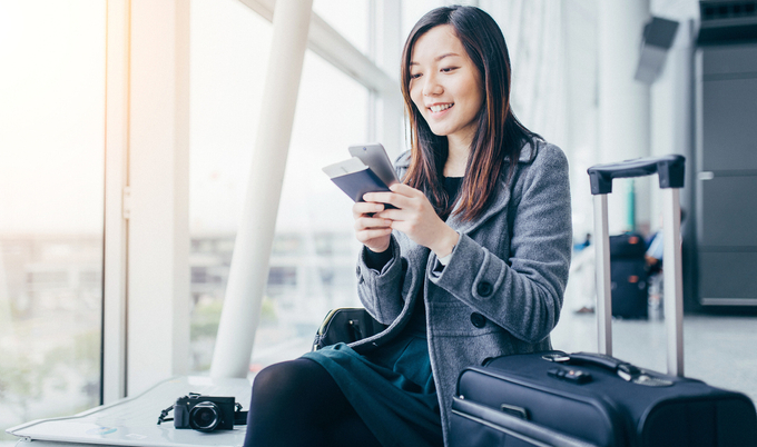 Young woman sitting at airport with her passport and phone in her hand, next to her luggage and a camera on the seat next to her.