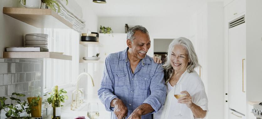 A smiling older couple is standing in a kitchen. The man is cutting vegetables on a cutting board. The woman has her hand on the man's shoulder.