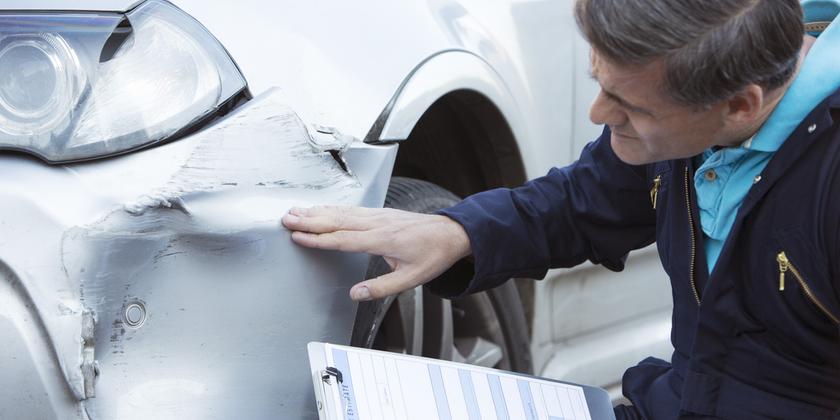 An auto workshop mechanic inspecting damage to the front driver side bumper of a car with a clip board in his hand.