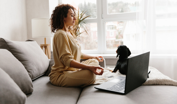 Woman meditating at home on a couch with a dog and laptop