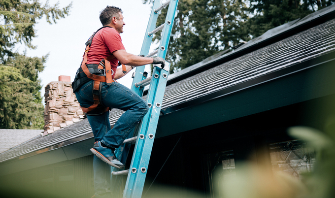 A professional male roofer is climbing up a blue ladder to get onto the roof of a home that has solar panels on it.