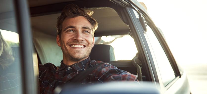 A smiling man with his seatbelt on is sitting in the driver's seat looking off into the distance.