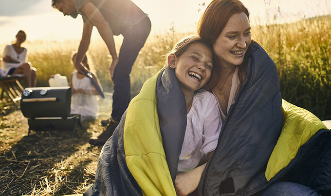Mom and daughter cuddled up under a sleeping bag with family in the background camping.