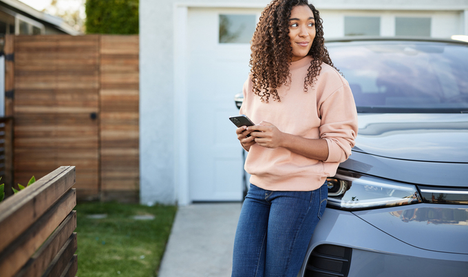 A woman in a sweatshirt and jeans is leaning on the front right corner of an electric car with a phone in her hands.