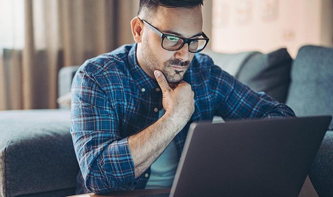 Man with glasses carefully looking at his laptop with a hand on his chin. 