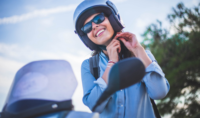 A woman is clipping the strap on a helmet as she is sitting on a moped.