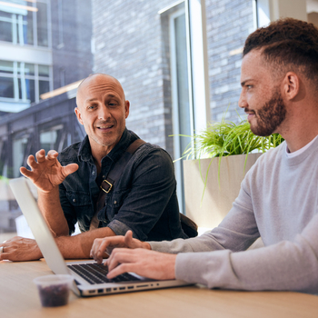 Two men discussing work at a laptop near a large window.