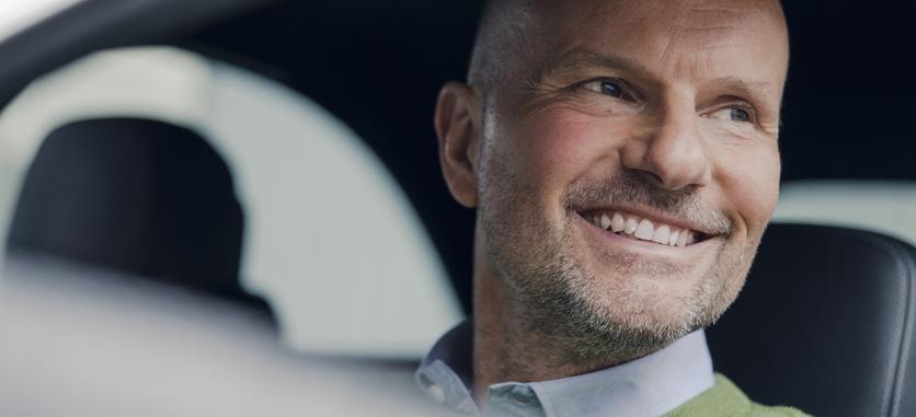 A close-up of a smiling older man sitting in the driver's seat of a car.