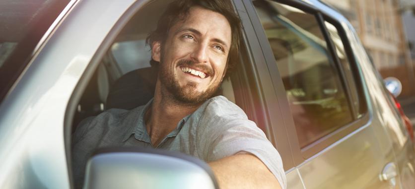 A smiling man looking out into the distance in a car with his arm over the car door.