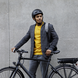 A young man in a helmet is standing behind a black eBike with a concrete wall in the background.