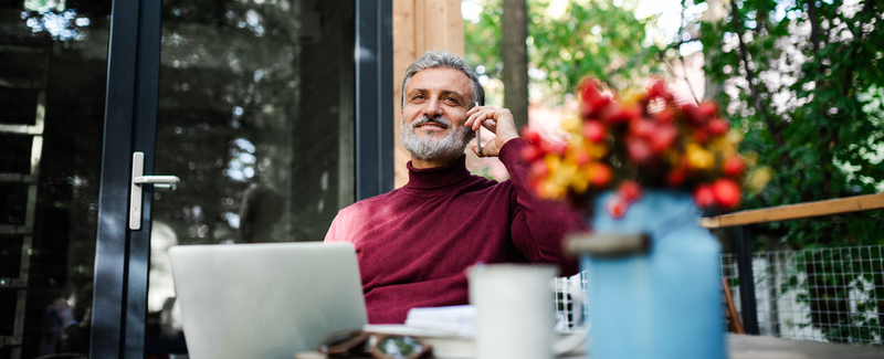 A mature man outdoors by a tree house, using laptop and smartphone.
