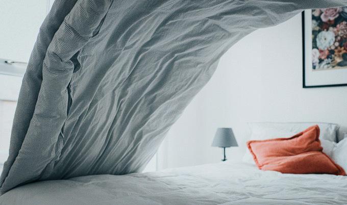 Gray bed comforter tossed in the air over a white bed.