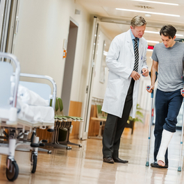 A doctor walking down the hall of a hospital with a patient on crutches.