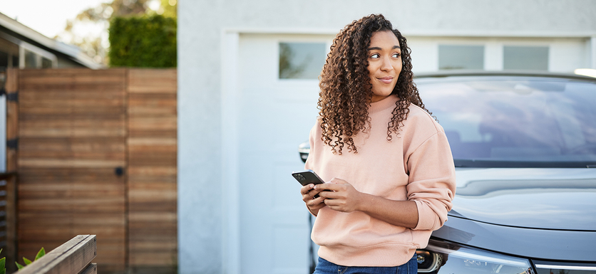 Contemplative woman with smart phone leaning on electric car parked in a driveway. 
