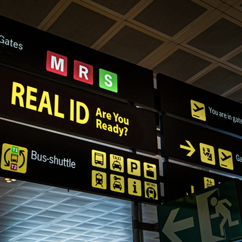 A digital display sign at an airport showing directional information that also says "REAL ID