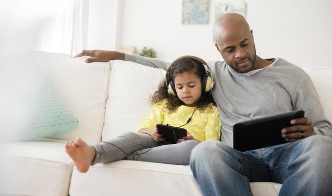 Dad and daughter sitting together on the couch, playing on electronic devices.