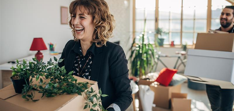 A smiling woman is holding a moving box and plant.