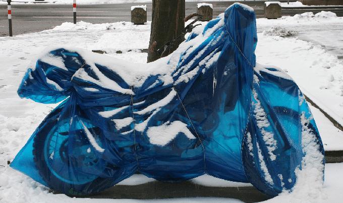 Motorcycle under a clear blue cover with snow on the ground.