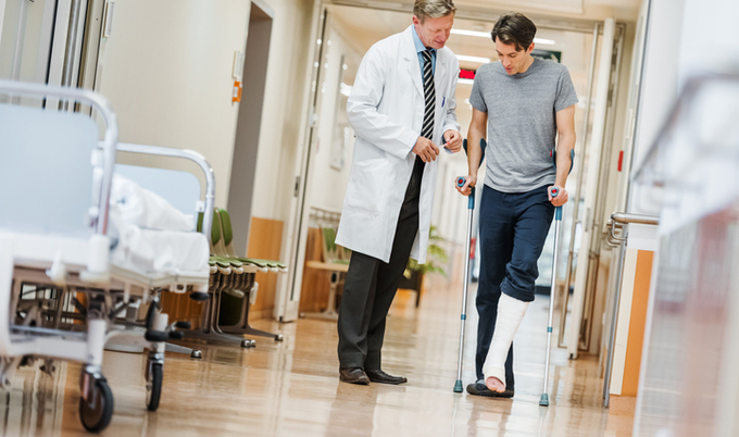 A doctor walking down the hall of a hospital with a patient on crutches.