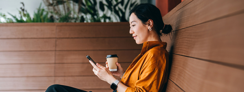 Young Asian woman sitting in a sidewalk cafe with a cup of coffee and using her smartphone. Enjoying a relaxing moment in the afternoon. 
