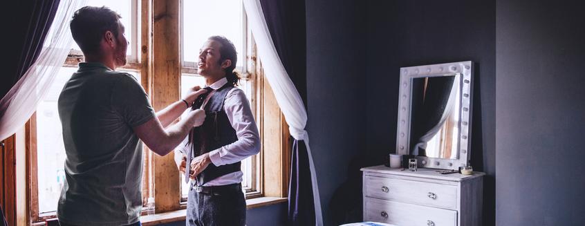 Two men&nbsp;are standing together in a bedroom while one is adjusting the&nbsp;other's tie.