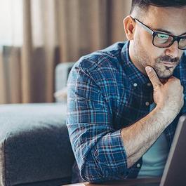 Man with glasses carefully looking at his laptop with a hand on his chin. 