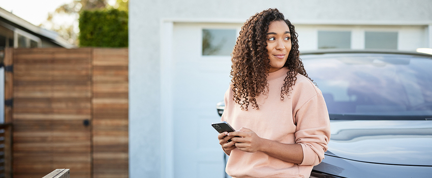Contemplative woman with smart phone leaning on electric car parked in a driveway. 