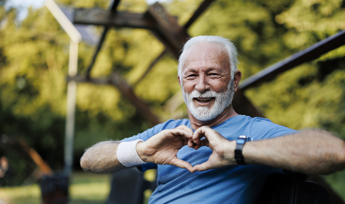 Older man sitting on a bench making a heart with his hands.