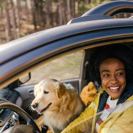 Young smiling woman driving a car with her dog is sitting in the passenger seat.