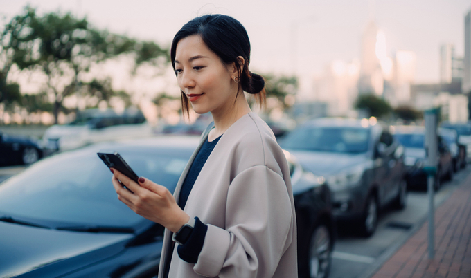A woman looking down at her phone with a street behind her that is lined with cars.